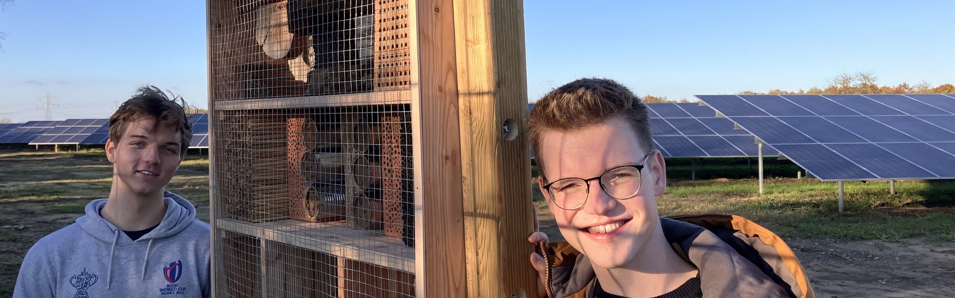 Zwei junge Erwachsene, die ihr Freiwilliges Ökologisches Jahr (FÖJ) bei Erdgas Südwest absolvieren, vor einem Wildbienenhabitat. Im Hintergrund ist eine Photovoltaik-Freiflächenanlage zu sehen.