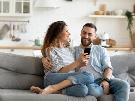 Ein junges Paar sitzt auf der Couch und ist glücklich. Die Frau zeigt dem Mann etwas auf dem Handy.