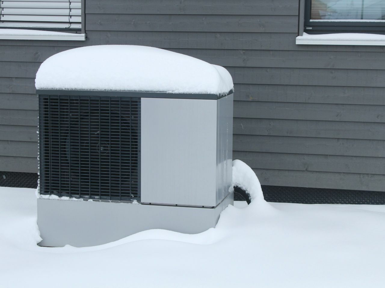 Eine Wärmepumpe steht vor einem Haus und ist mit Schnee bedeckt. Die Wärmepumpe funktioniert auch im Winter effektiv.