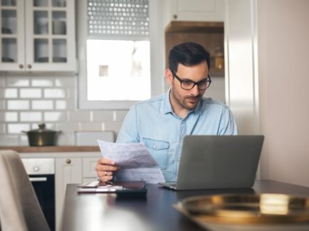 Ein Mann im Hemd sitzt am Esstisch in seiner Küche vor seinem Laptop und blickt freundlich auf den Bildschirm, während er seine Rechnung in der Hand hält.