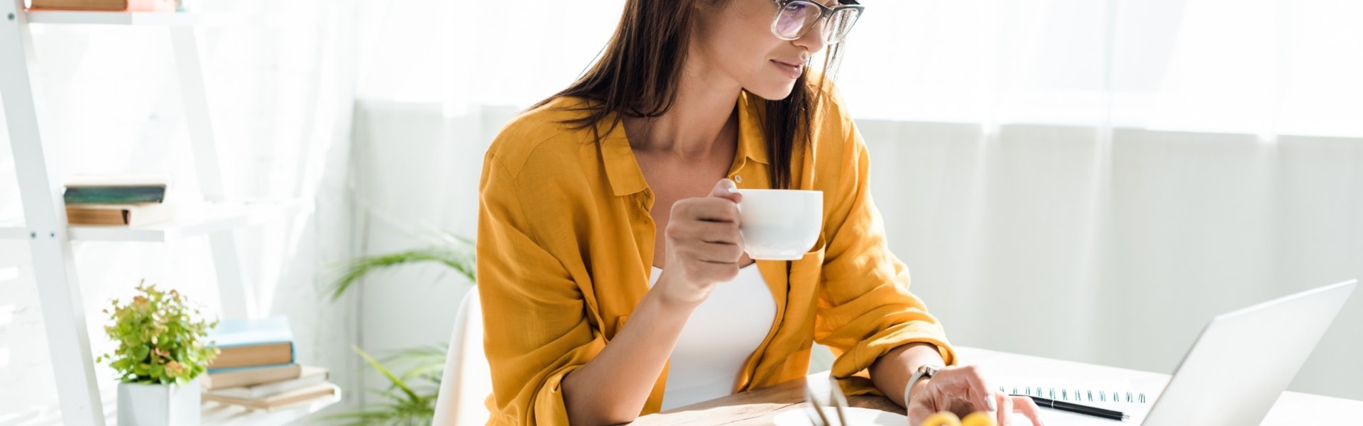 Eine Frau sitzt in einem hellen Büro an einem Schreibtisch vor ihrem Laptop, Notizen und Büromaterial, in der Hand eine Tasse Kaffee.