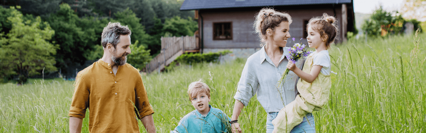 Eine Familie steht auf ihrem großen Grundstück vor ihrem Zuhause mit PV-Anlage auf dem Dach. Die Familie hält sich dabei an den Händen und wirkt glücklich.